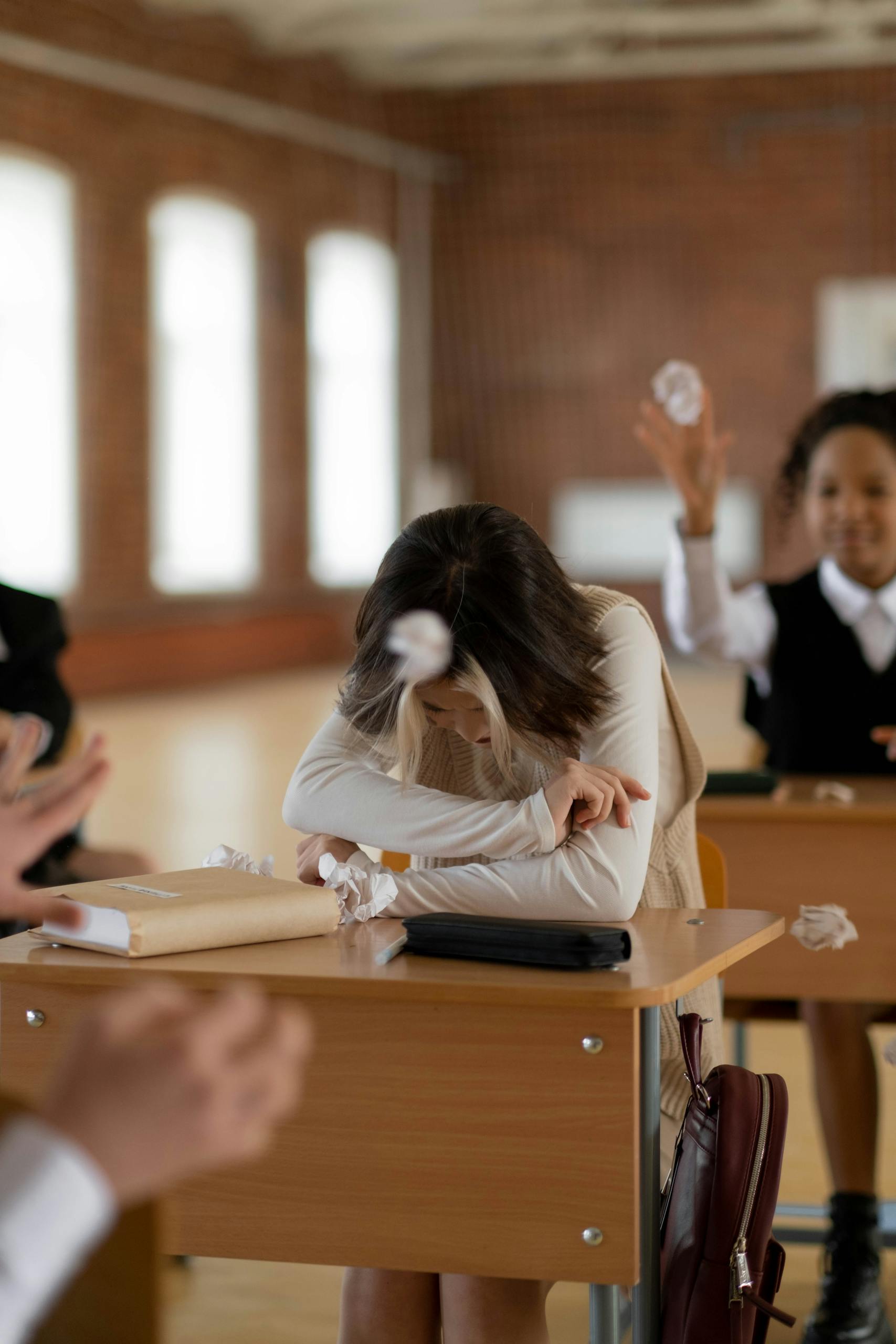 A teenage girl sits at her desk, visibly upset as classmates throw paper balls at her.