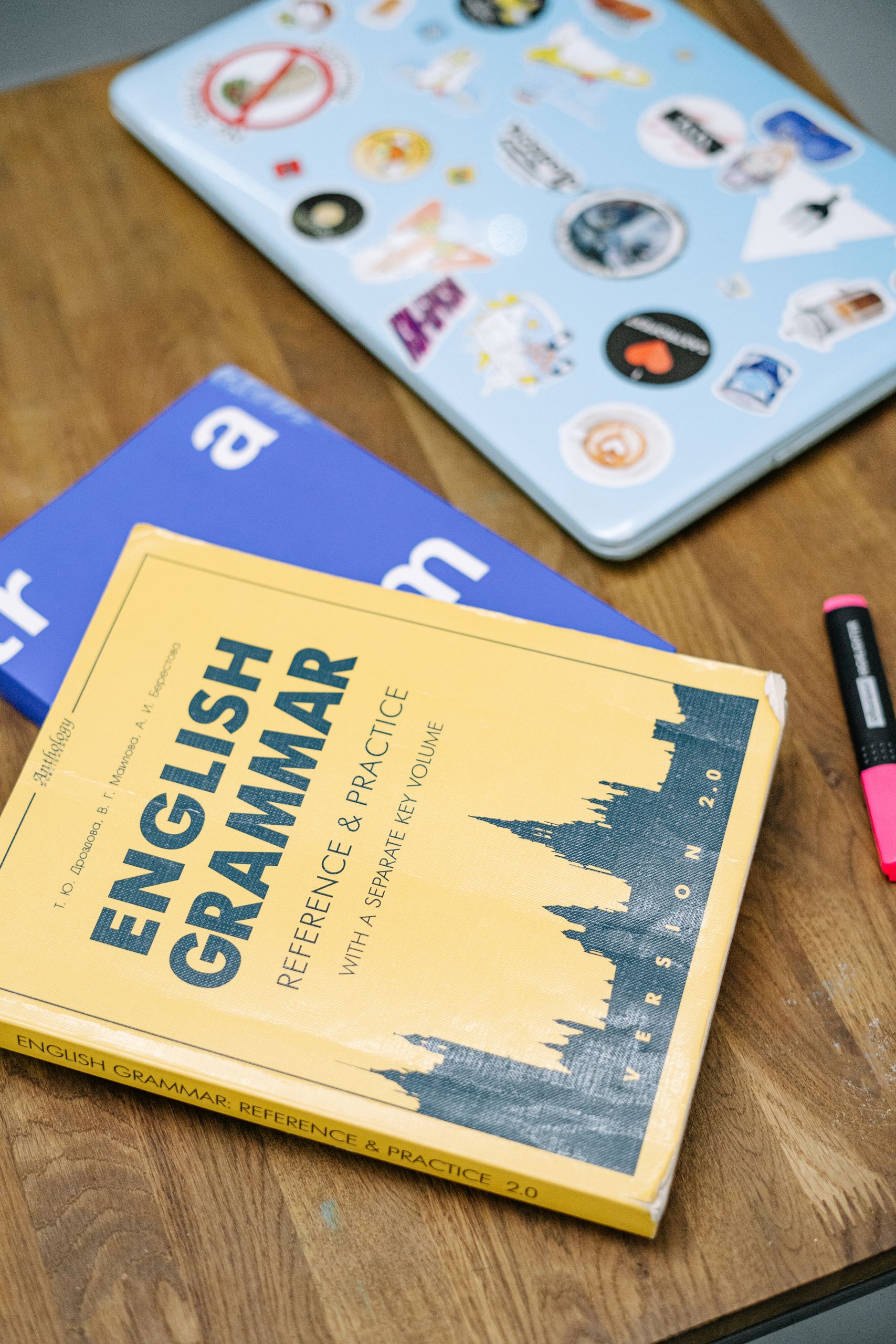 High-angle view of an English grammar book and a laptop with stickers on a wooden desk.